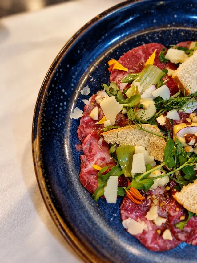 Beef carpaccio on a blue speckled plate with rocket, shaved cheese, croutons and orange petals
