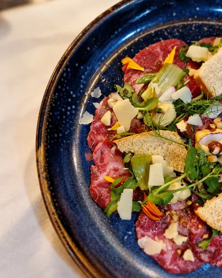 Beef carpaccio on a blue speckled plate with rocket, shaved cheese, croutons and orange petals