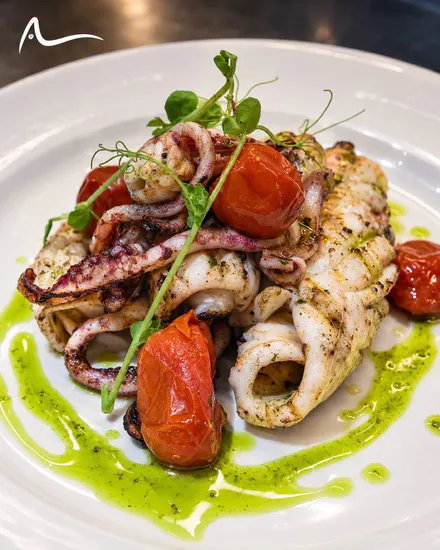 Grilled squid bodies and tentacles with blistered cherry tomatoes and pea shoots, inside a ring of green herb oil on a white plate