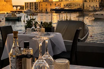 A terrace table set for dinner under a parasol, the harbour behind it at sunset