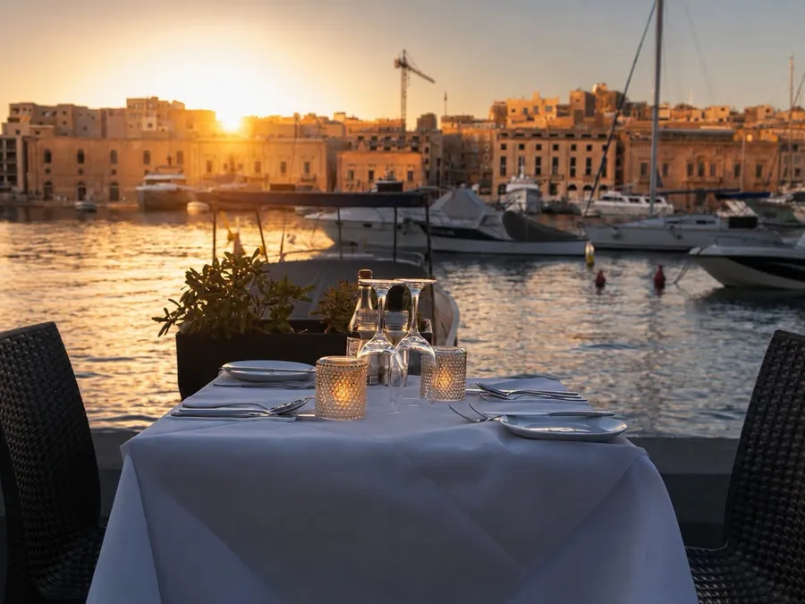 A laid terrace table with candles and glassware, the sun setting over the harbour behind it