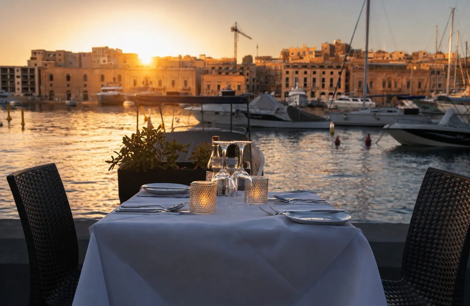 A laid table at the water’s edge at sunset with boats behind